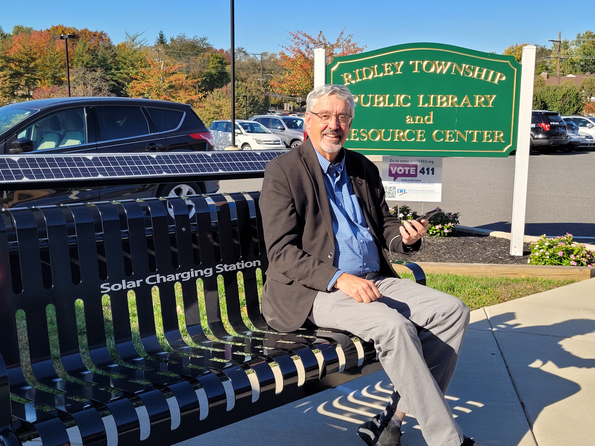 Senator Tim Kearney sitting on a solar bench with the Ridley Township Public library and Resource Center sign in the background.